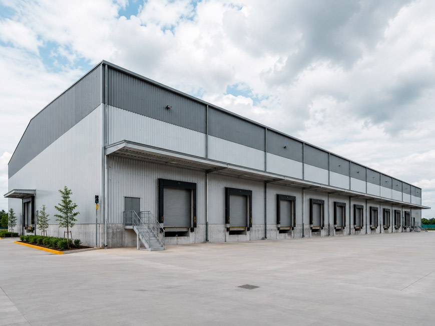 Fotografía exterior de una nave industrial ligera tipo bodega, estructura metálica prefabricada con lámina galvanizada en tonos grises, andenes de carga con cortinas metálicas, amplio patio de maniobras de concreto, cielo nublado claro, perspectiva de tres cuartos que muestra la longitud del edificio, estilo fotografía industrial corporativa, sin vehículos de marca visibles, sin texto ni logotipos.